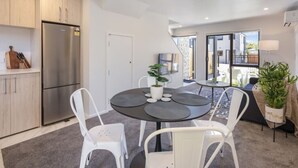 A modern kitchen with a stainless steel refrigerator, white cabinets, and a dining area with a round table and white chairs. - Ariki Shire: A Wellington Fantasy (Lower Hutt)
