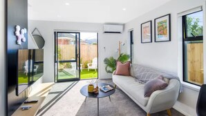 A modern living room with a sofa, coffee table, and a TV. There is a sliding glass door leading to a backyard with a lawn and chairs. - Staycation Treasure at Johnston (Lower Hutt)