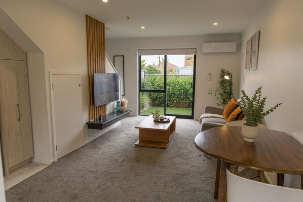 A modern living room with a flat-screen TV, a wooden coffee table, a sofa, and a dining table with a potted plant.