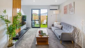 Una sala de estar moderna con un sofá gris, una mesita de centro y un televisor sobre un mueble. Hay una puerta corrediza de vidrio que da a un patio trasero con una silla blanca y una valla de madera.