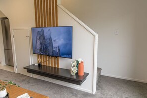 A flat-screen TV mounted on a wooden wall, a small table with a vase and a plant, and a staircase with a white railing. - Hideaway in the Grove (Lower Hutt)