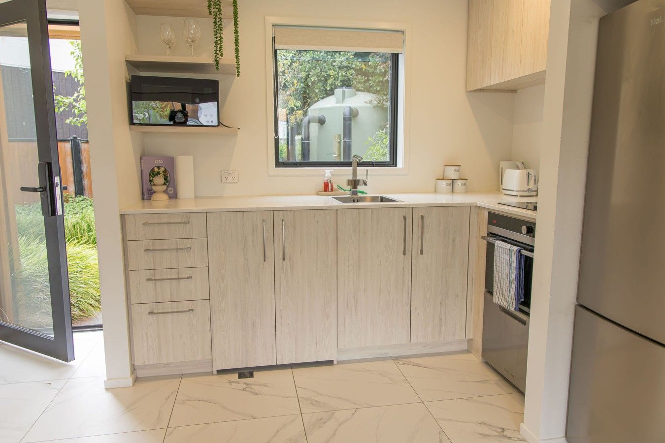 A modern kitchen with a built-in oven, a stainless steel sink, and a window overlooking a garden.