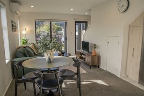 A modern living room with a round dining table, a sofa, a TV on a wooden cabinet, and a large window.