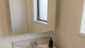 A bathroom with a white marble sink, a mirror above it, and a small window. - Retreat at Biddle Crescent (Lower Hutt)