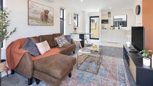 A modern living room with a brown leather sofa, a coffee table, and a TV. There is a kitchen area in the background with white cabinets and a dining table. - Radiant Spot on Biddle Crescent (Lower Hutt)