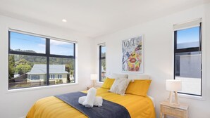 A modern bedroom with a large bed, two bedside tables, and a window with a view of a house and trees. - Radiant Spot on Biddle Crescent (Lower Hutt)