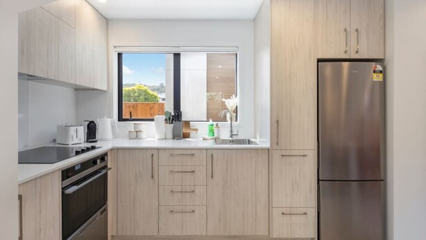 A modern kitchen with light wood cabinets, stainless steel appliances, and a large window offering a view of greenery. - Biddle Cres Elegance (Lower Hutt)