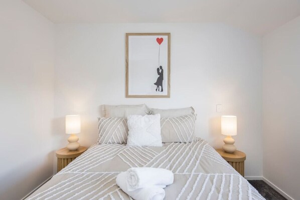 Una cama bien hecha con ropa de cama a rayas, dos lámparas de noche y un cuadro en la pared de una persona sosteniendo un corazón.