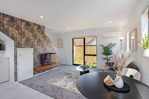 A modern living room with a TV, a fireplace, a dining area with a table, and a large window. - Cosy Retreat at Biddle Crescent (Lower Hutt)