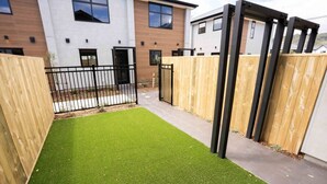 A courtyard with artificial green turf, wooden fences, and a black metal gate. - The Biddle Haven: 2BR Retreat (Lower Hutt)
