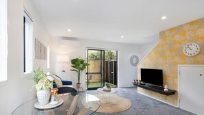 Un salon moderne avec une table basse en verre, un canapé, un téléviseur fixé au mur, qui est décoré d’un motif jaune, et une porte-fenêtre coulissante donnant sur un jardin.