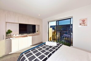 Een moderne slaapkamer met een groot bed, een televisie op een inbouwplank, een balkon met uitzicht over de stad en een kleine plant op de plank.