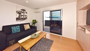 A modern living room with a sofa, a coffee table, a TV, and a sliding glass door leading to an outdoor area. - Spacious CBD 1-Bed 1-Bath (Christchurch)