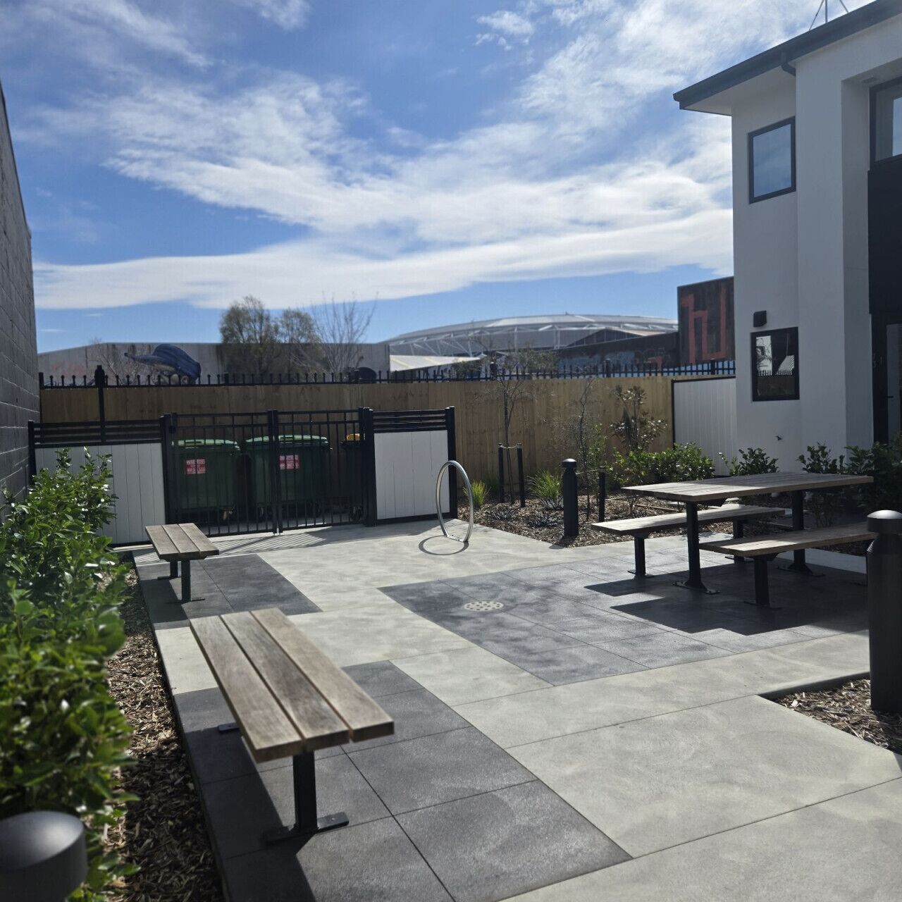 En udendørs patio med beton gulv, træbænke og en metalring struktur.