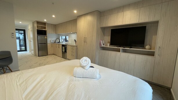 Une chambre d’hôtel moderne dotée d’un grand lit, d’un téléviseur fixé au mur, d’une kitchenette et d’une salle de bain visible à travers une porte ouverte.