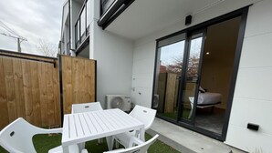 Une terrasse avec une table et des chaises blanches, une clôture en bois et une porte-fenêtre coulissante donnant sur une pièce où se trouve un lit.