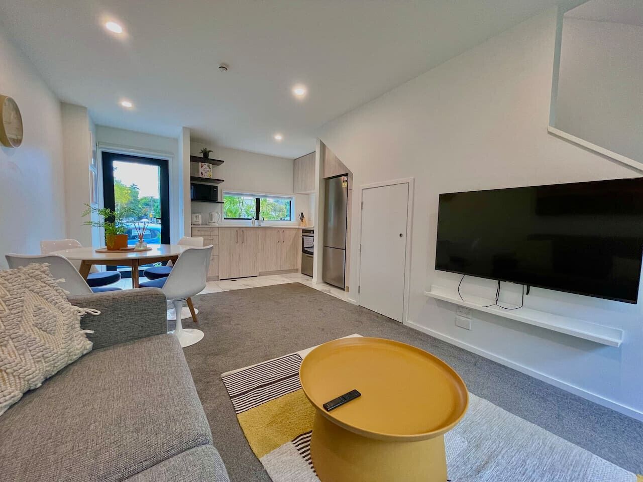 Een moderne woonkamer met een bank, een salontafel en een televisie. Achtergrond is een keuken met kasten en een koelkast.