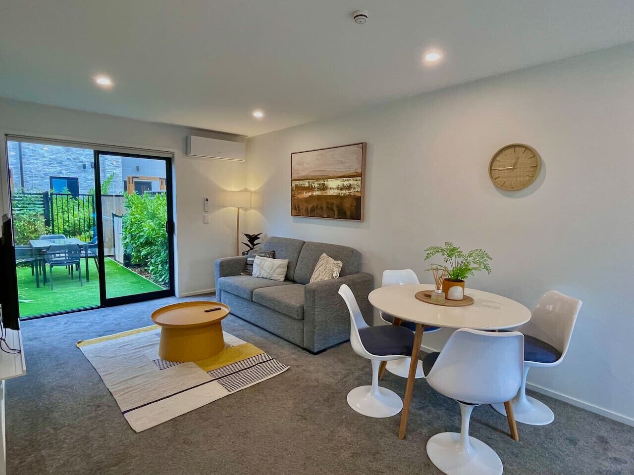 Een moderne woonkamer met een grijze bank, een ronde salontafel en een eettafel met stoelen. Er is een groot raam met uitzicht op een tuin en een klok aan de muur.