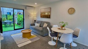 Un salon moderne avec un canapé gris, une table basse ronde et une table à manger avec des chaises. Une grande fenêtre offre une vue sur un jardin, et une horloge est accrochée au mur.