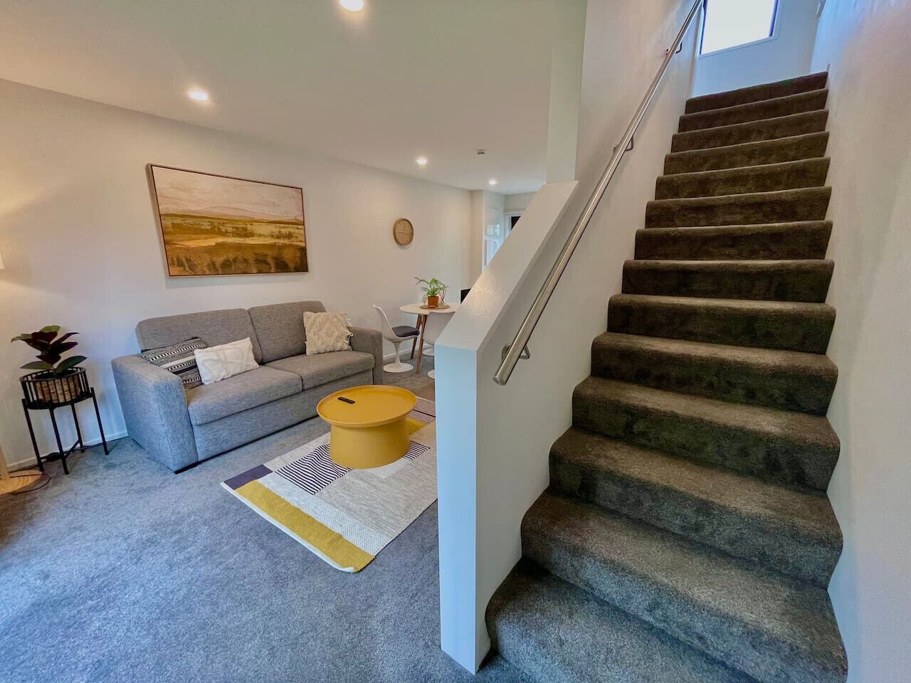 Een moderne woonkamer met een grijze bank, een gele poef en een trap naar een verdieping erboven.