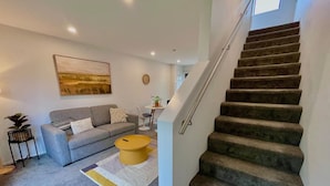 Un salon moderne avec un canapé gris, un pouf jaune et un escalier menant à l’étage supérieur.