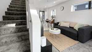 Una sala de estar moderna con una escalera, un sofá negro, una mesa de centro blanca y un ventanal grande.