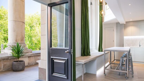 A modern kitchen with a built-in bench and stools, a large window with green curtains, and a potted plant outside.