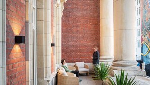 A lobby with brick walls, a mural, and a column. There are wicker chairs and a potted plant.