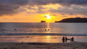 Escena de playa con gente nadando y relajándose cerca de la orilla al atardecer.