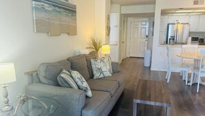 A living room with a grey sofa, a glass coffee table, and a painting of a beach scene. - 2-bedroom condo in gorgeous North Topsail Beach with WiFi, AC viewing the ocean (North Topsail Beach)