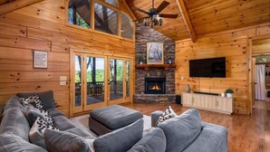 A log cabin interior with a stone fireplace, a large gray sofa, and a television mounted on the wall. - Newly renovated  5BR 3BA Cabin with amazing Mountain Sunset views (Morganton)