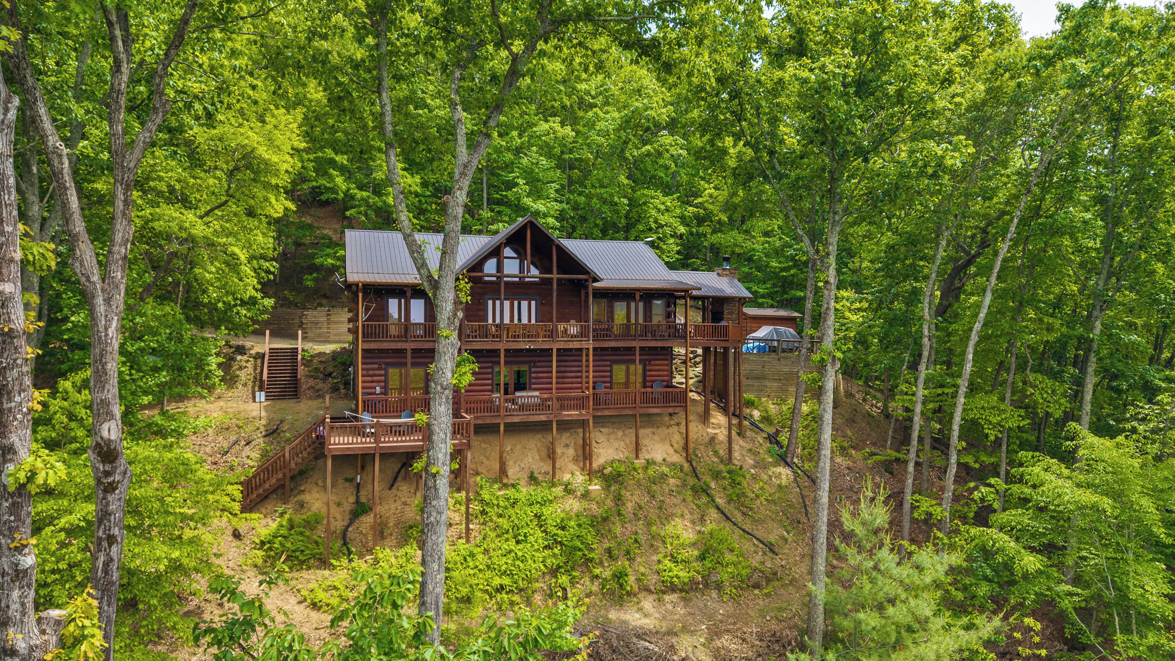 A two-story log cabin with a deck, surrounded by dense forest.