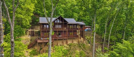 A two-story log cabin with a deck, surrounded by dense forest.