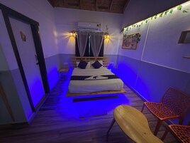 Una habitación pequeña y acogedora con cama, escritorio y silla. Hay una ventana con cortinas, un equipo de aire acondicionado y un estante en la pared.