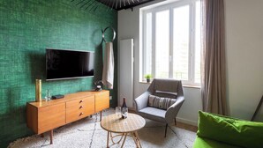 A living room with a green textured wall, a flat-screen TV, a wooden cabinet, a round wicker table, a grey armchair, and a green sofa. - Dify Art Deco - Charpennes (Villeurbanne)