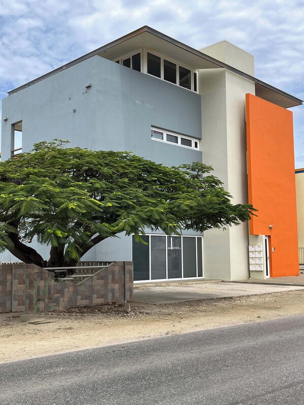 Un edificio moderno con un árbol grande en primer plano, una entrada con portones y un cielo despejado.