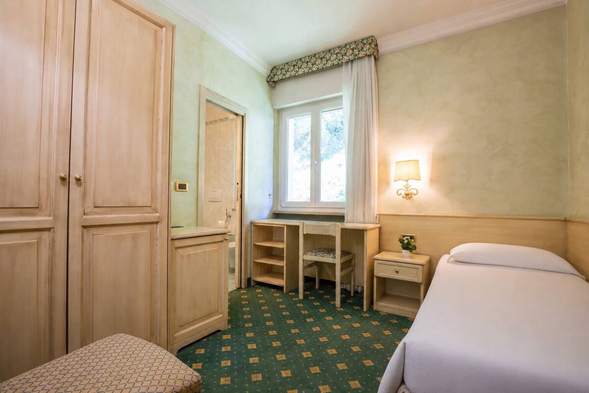 Una camera d'albergo con un letto, una scrivania, una sedia, un tavolino, un comodino, una lampada e una finestra con tende.