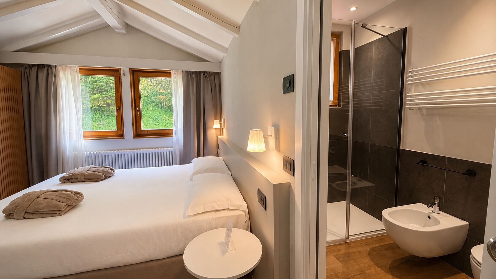 A modern hotel room with a large bed, a round bedside table, a glass shower enclosure, and a separate bathroom with a sink and toilet.