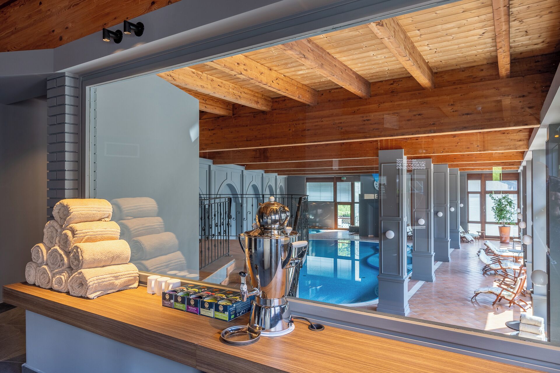 Une chambre d’hôtel avec un plafond en bois, un grand miroir reflétant l’espace piscine, et un comptoir avec des serviettes et des tasses à thé.