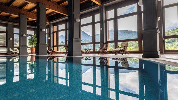 Una amplia piscina interior con reflejos cristalinos, vigas de madera y grandes ventanales que ofrecen vistas a la montaña.