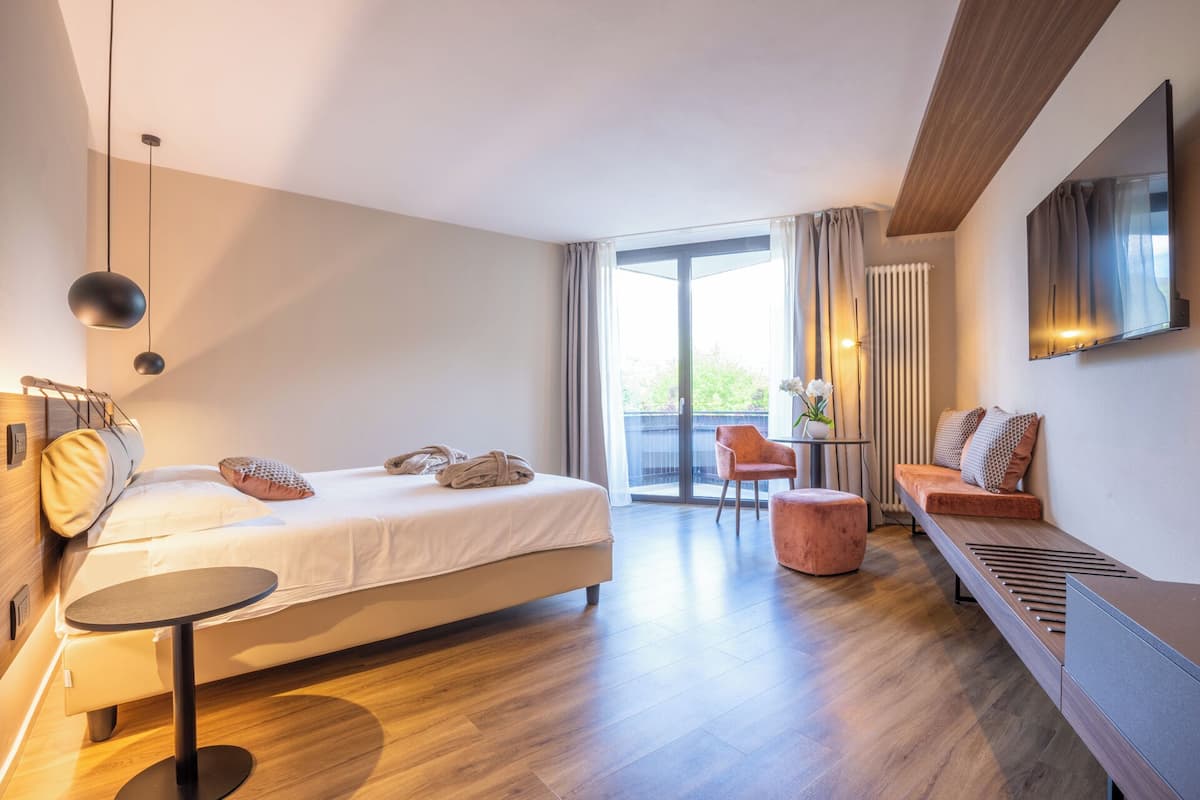 Una moderna camera d'albergo con un letto, un piccolo tavolino rotondo, una poltrona e una grande finestra con tende.