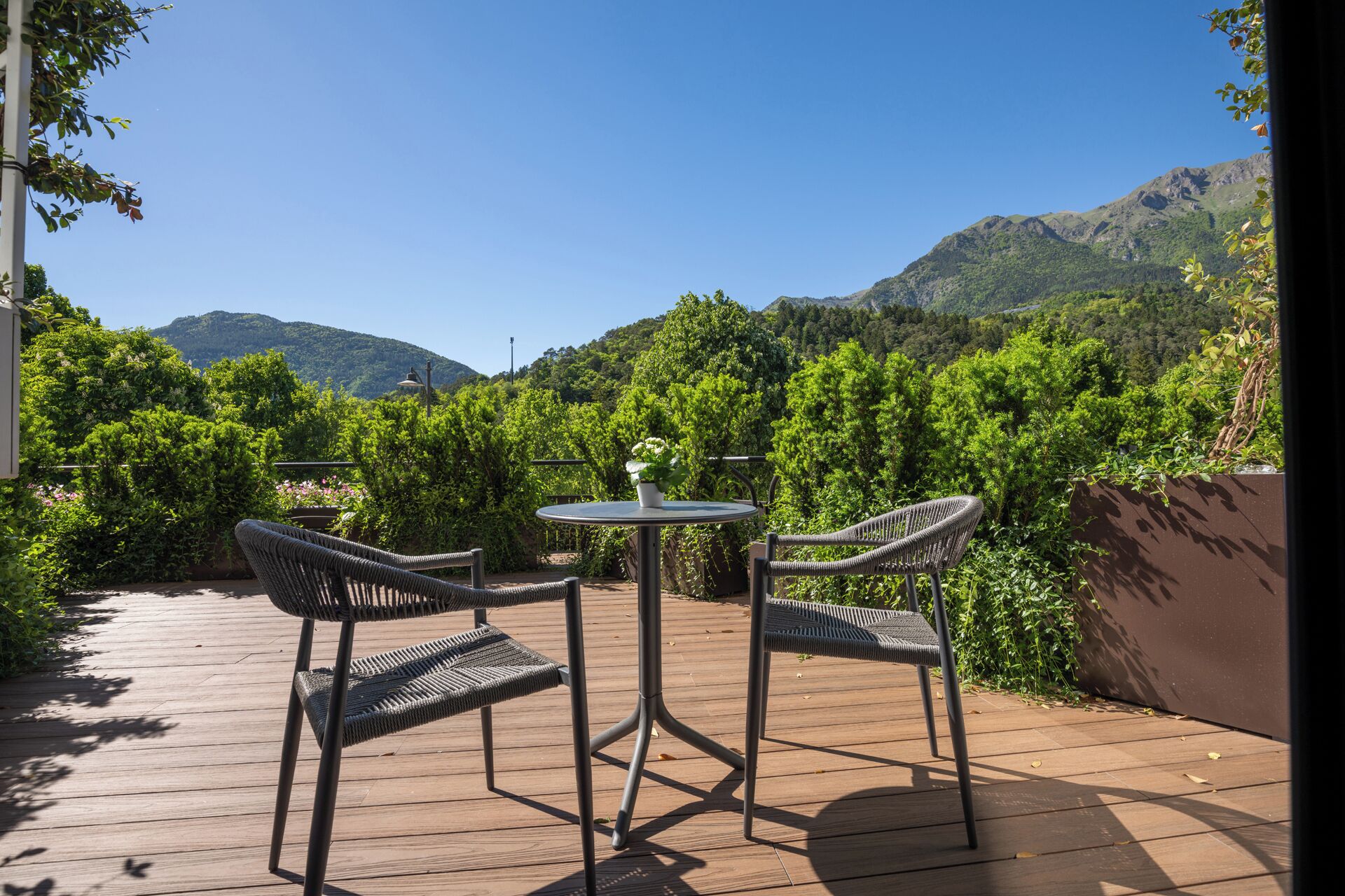 An outdoor patio with wicker chairs and a table, surrounded by lush greenery and mountains in the background.
