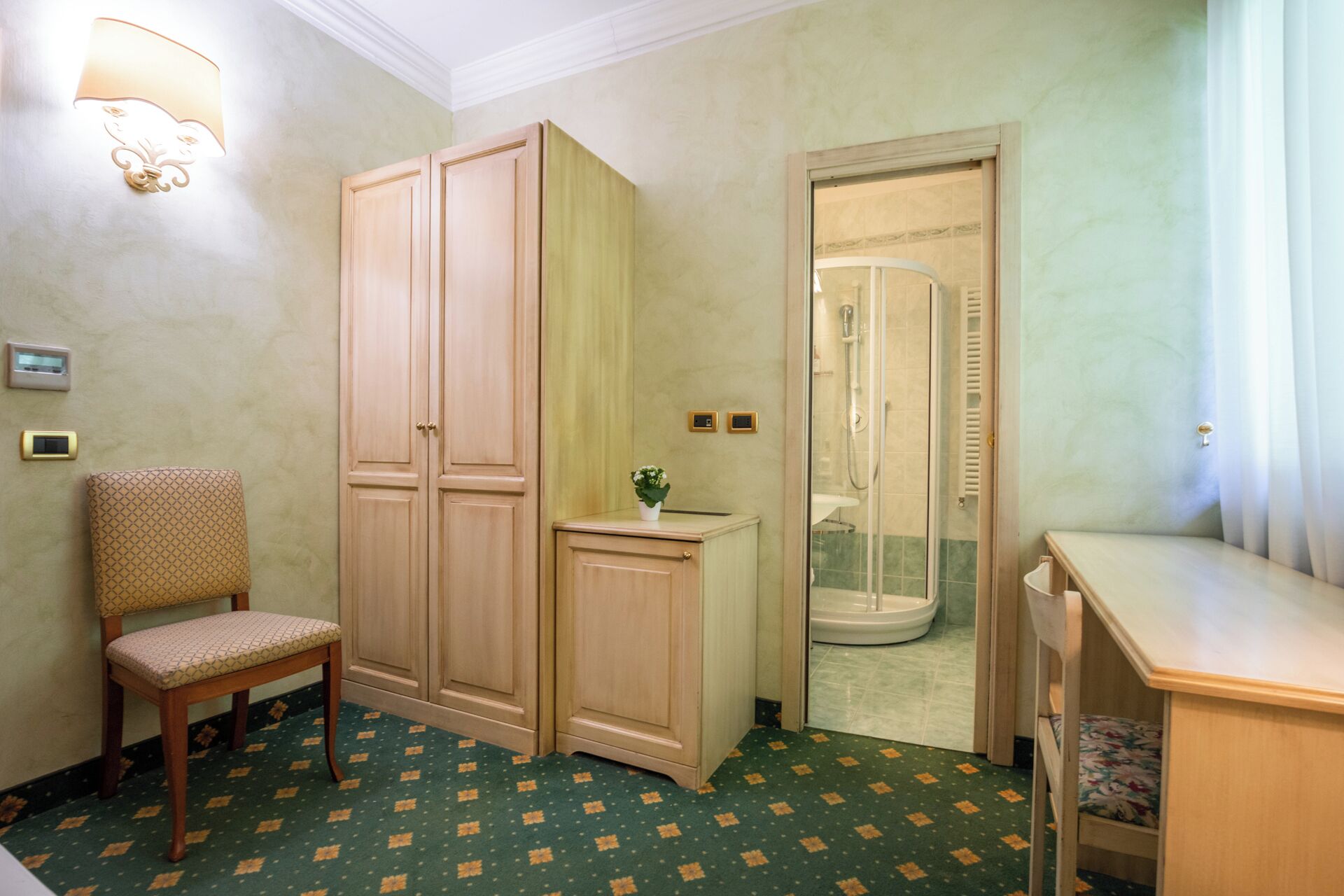 A hotel room with a wooden wardrobe, a chair, a small table, a shower area, and a green patterned carpet.