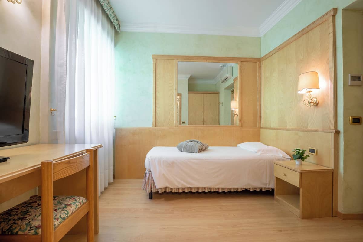 Una camera d'albergo con un letto, un comodino, uno specchio, una TV e una finestra con tende.