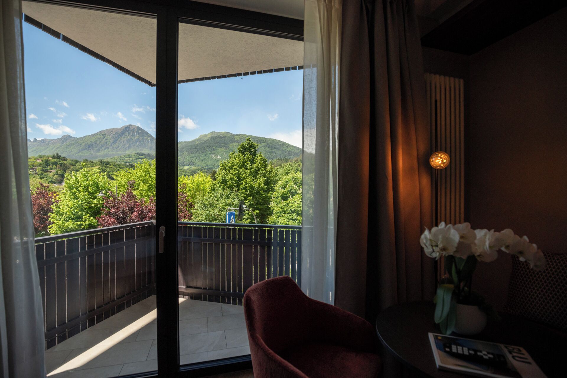 A balcony with a view of mountains and greenery, a red chair, a table with a vase of flowers, and a remote control.