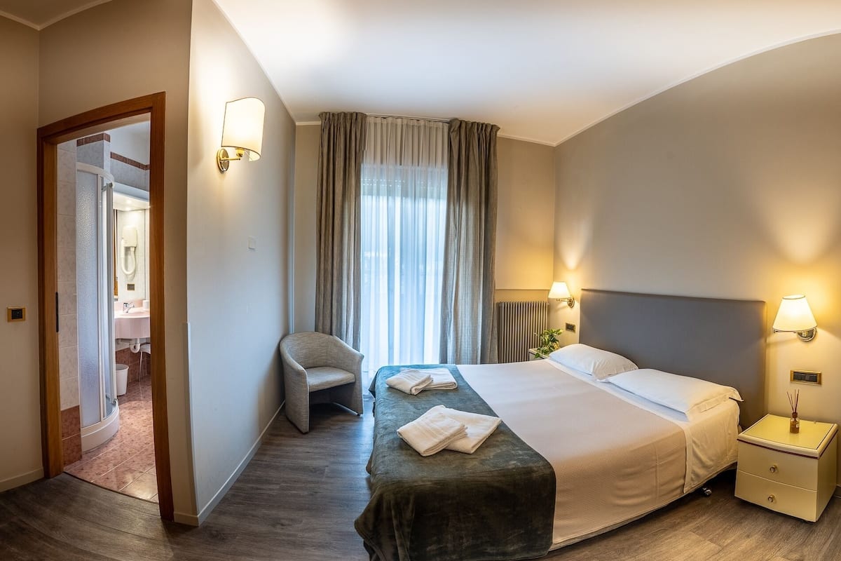 Una camera d'albergo con un letto, una sedia, un comodino, una lampada e vista sulla città dalla finestra.