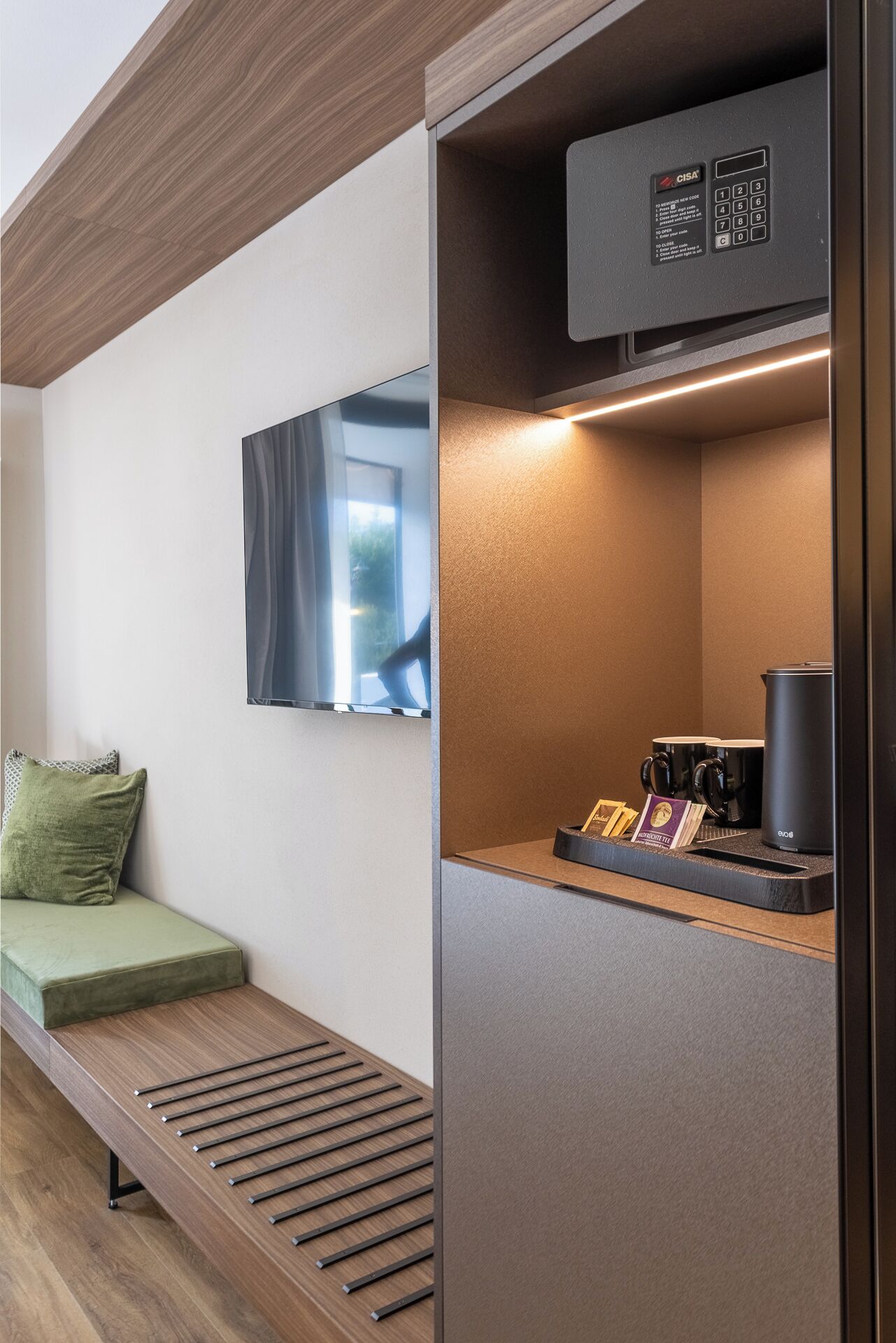 A modern hotel room with a wooden bench, a flat-screen TV mounted on the wall, a coffee station with mugs and a kettle, and a GISA charging station.