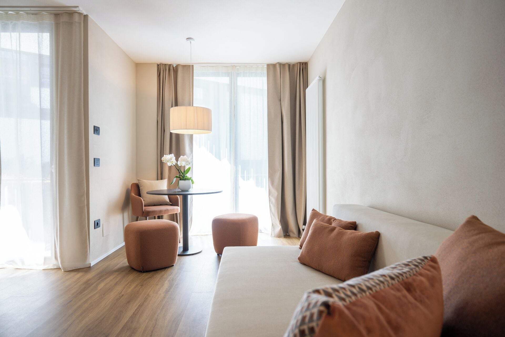A hotel room with a bed, a sofa, a round table with a lamp, a chair, and a potted plant.