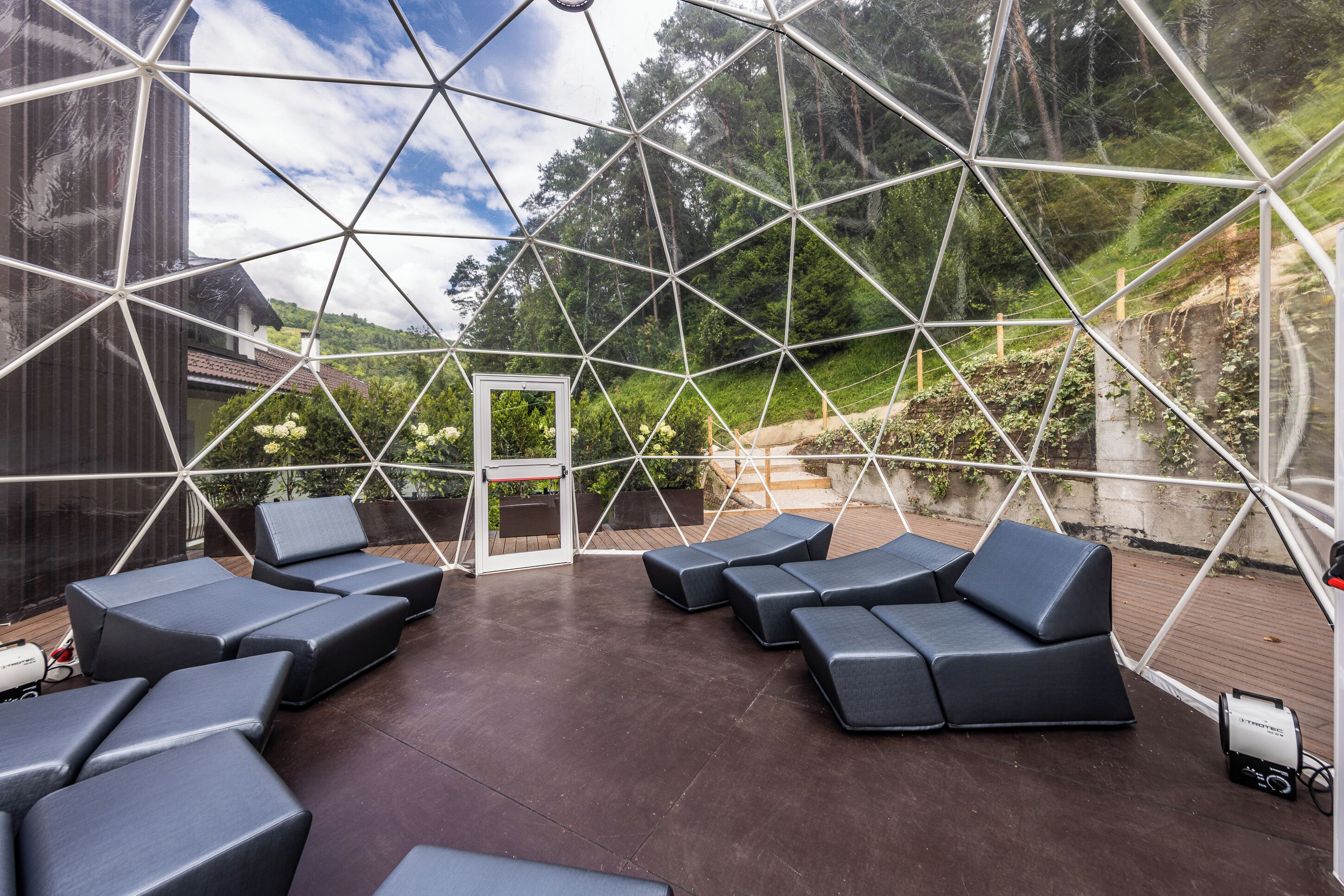 A geodesic dome structure with a patio area featuring multiple seating options and a view of a forested hillside.