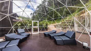 A geodesic dome structure with a patio area featuring multiple seating options and a view of a forested hillside.